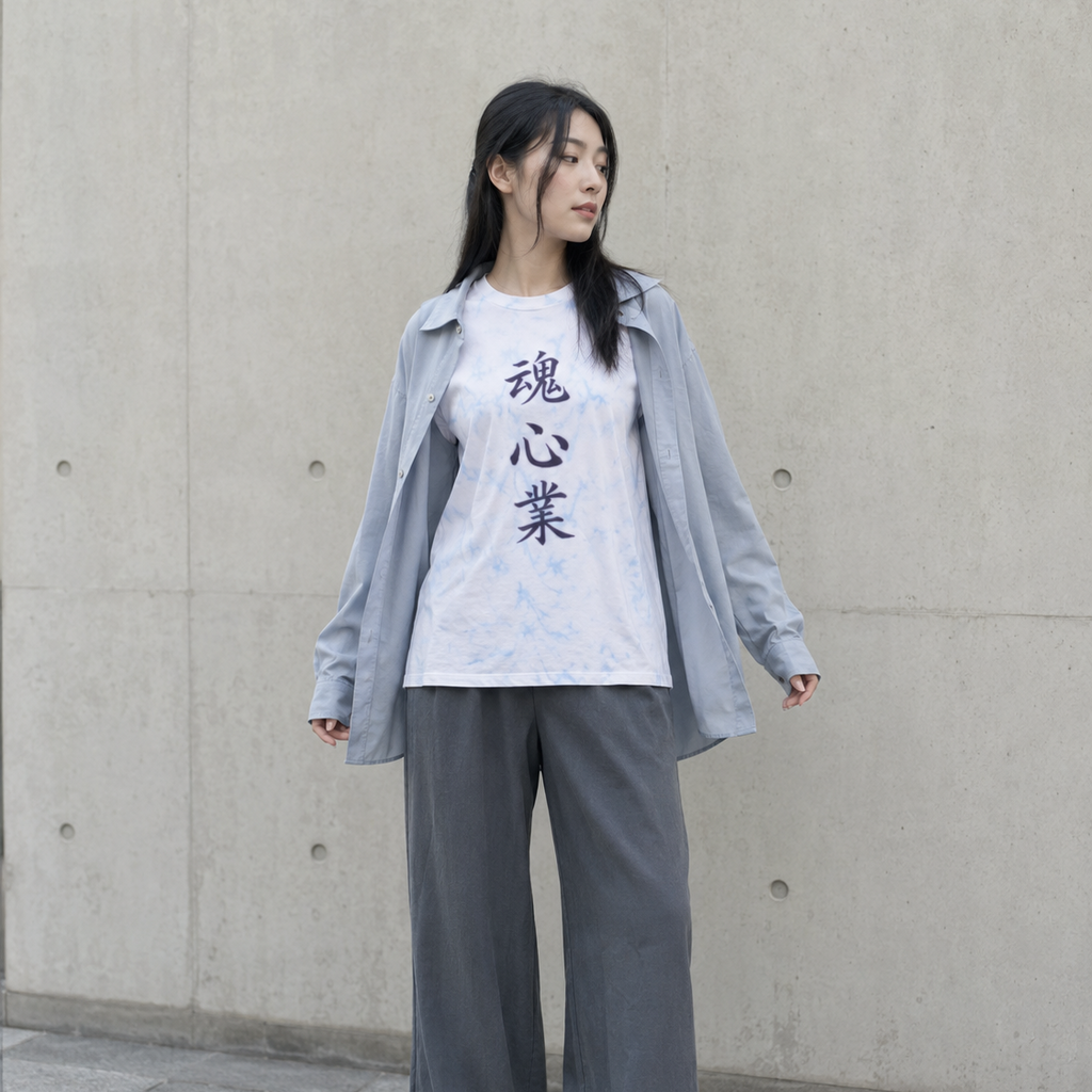 A woman stands against a concrete wall wearing a white graphic T-shirt with black Japanese characters, a light blue button-up shirt layered over it, loose gray wide-leg pants, and white sneakers.