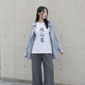 A woman stands against a concrete wall wearing a white graphic T-shirt with black Japanese characters, a light blue button-up shirt layered over it, loose gray wide-leg pants, and white sneakers.
