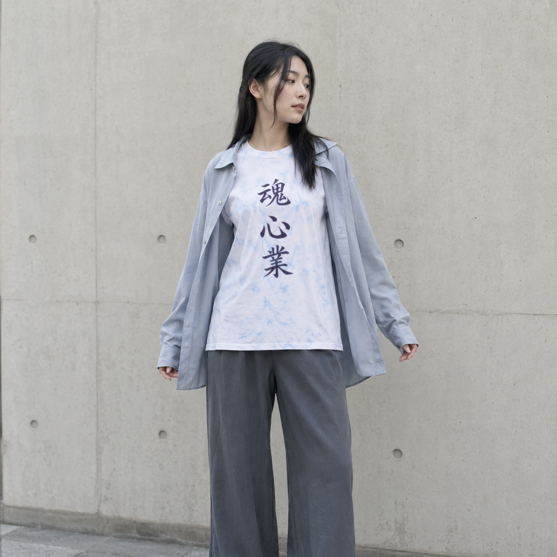 A woman stands against a concrete wall wearing a white graphic T-shirt with black Japanese characters, a light blue button-up shirt layered over it, loose gray wide-leg pants, and white sneakers.
