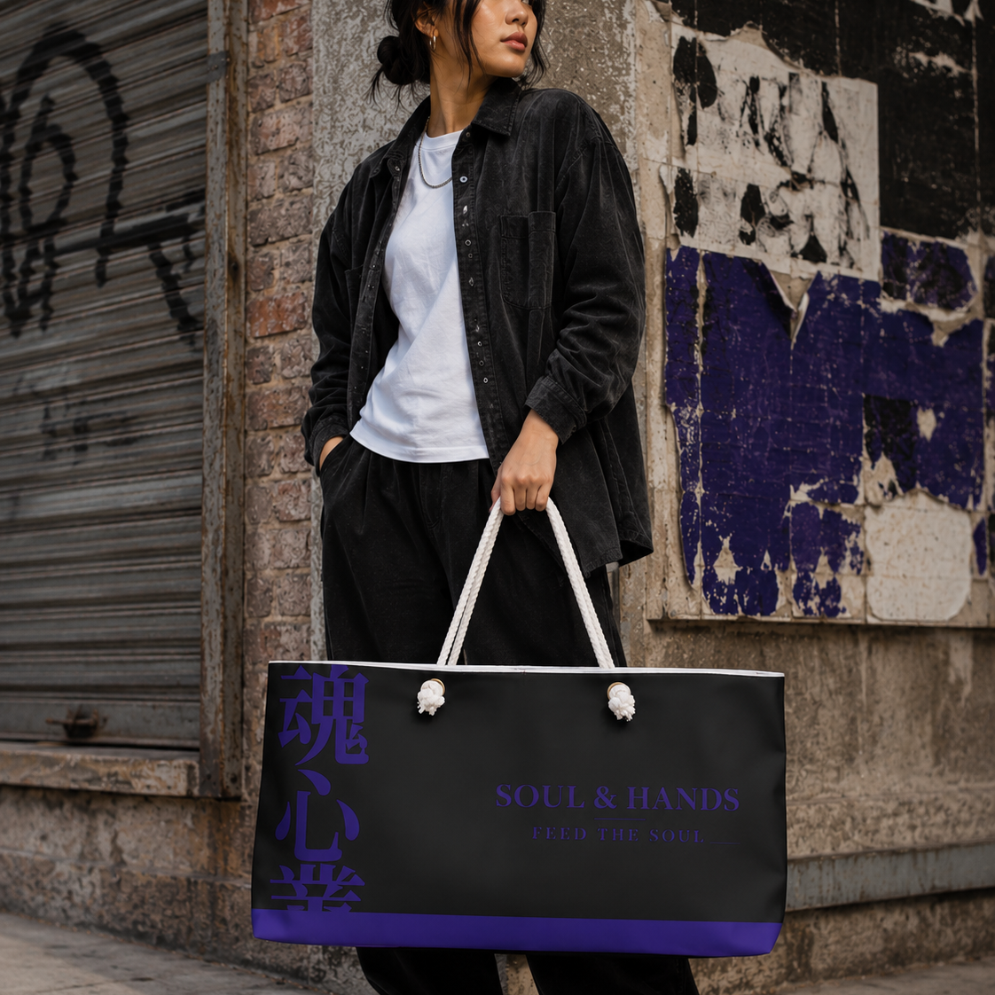 Woman carrying a black Soul & Hands weekender bag with purple trim, Japanese kanji and Feed the Soul branding.