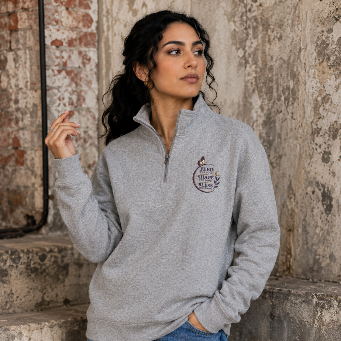 Woman wearing a grey Soul & Hands quarter-zip sweatshirt with a purple butterfly and Feed, Shape, Bless chest design.