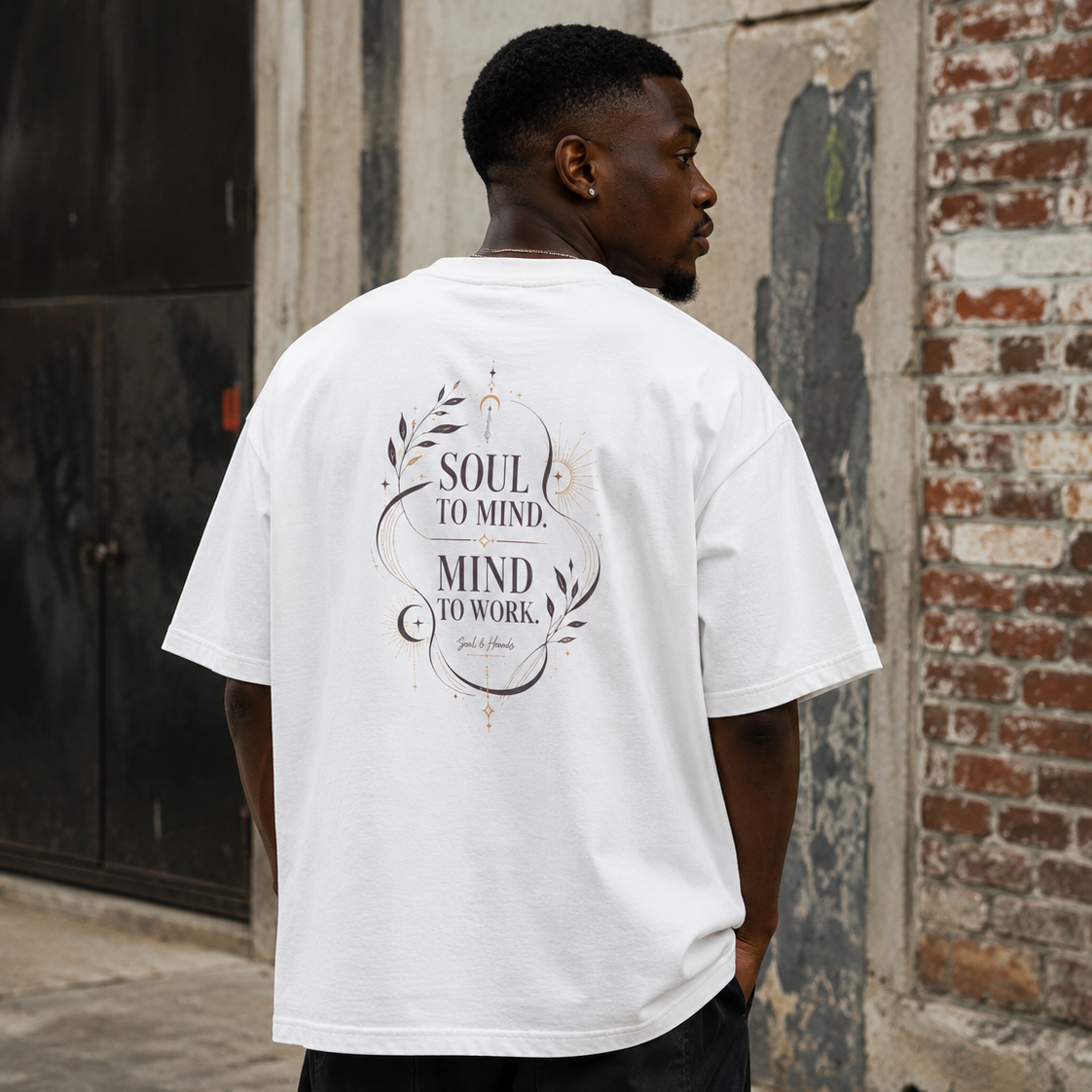 Back view of a man wearing a white Soul & Hands oversized T-shirt with black-and-gold Soul to Mind, Mind to Work botanical artwork.