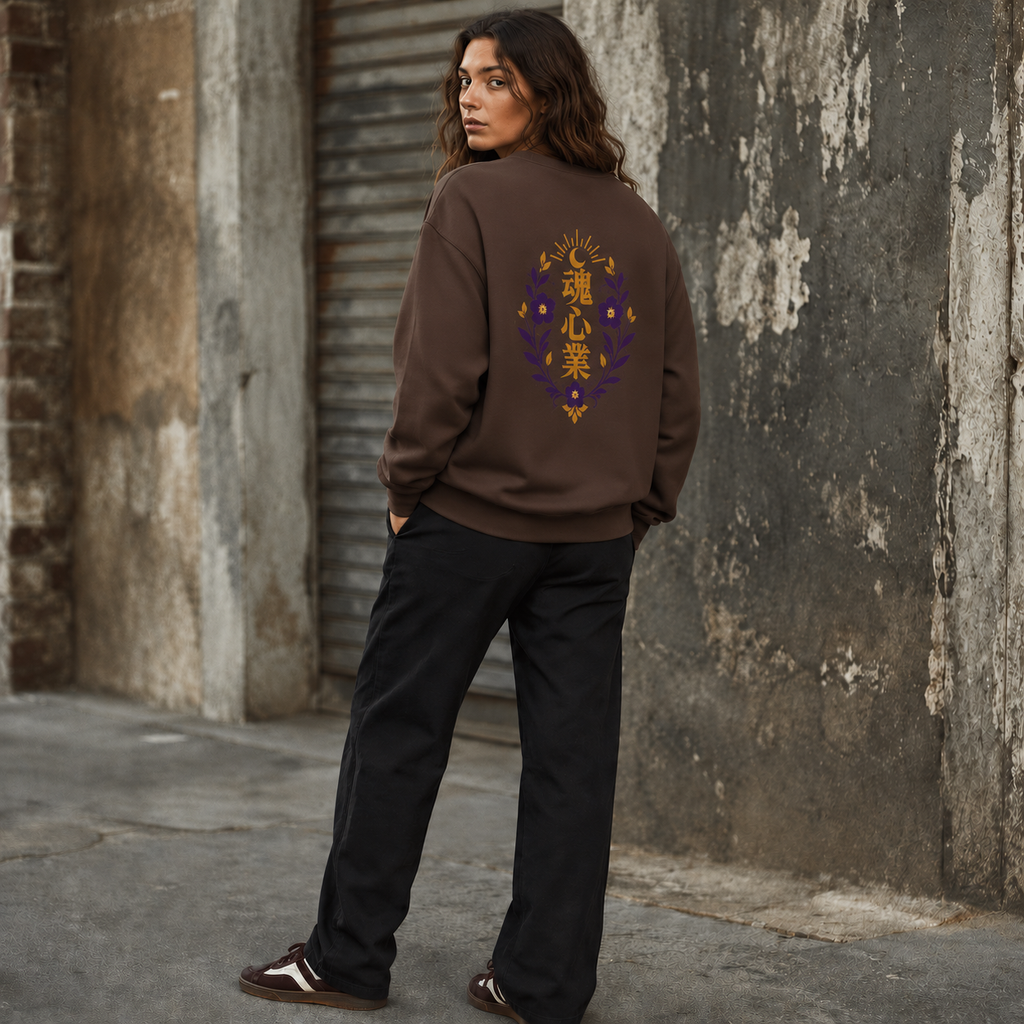 Woman wearing a plain brown crewneck sweatshirt with a gold and purple Japanese kanji floral crest on the back, styled with black wide-leg trousers in an urban setting.