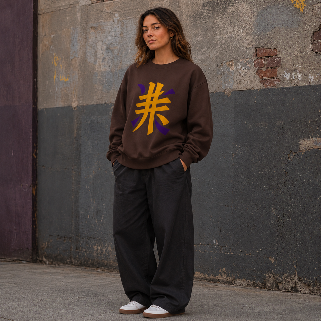 Woman wearing a plain brown crewneck sweatshirt with a bold gold and purple Japanese kanji design on the front, styled with black wide-leg trousers in an urban setting.