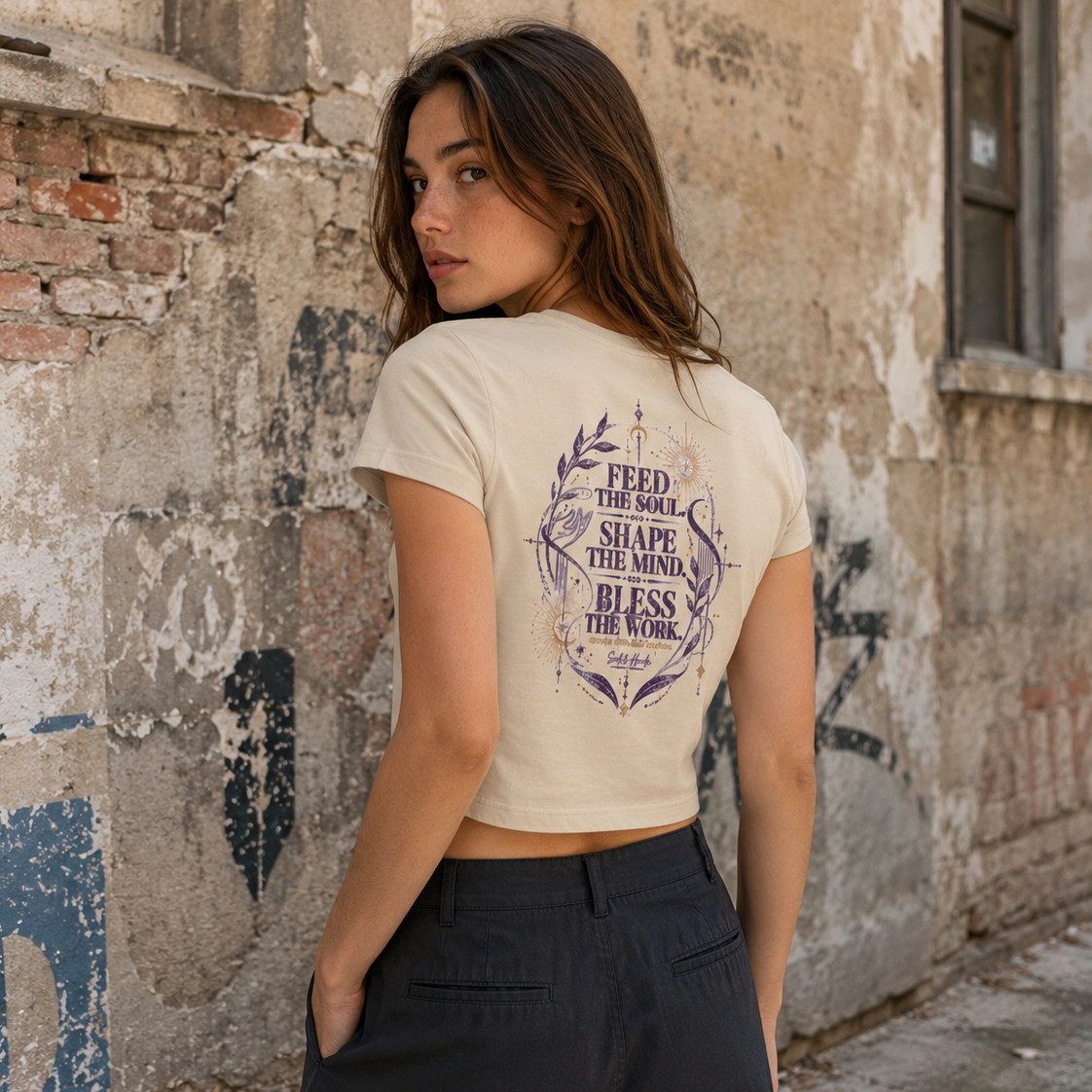 Back view of a woman wearing a sand Soul & Hands crop T-shirt with purple Feed the Soul, Shape the Mind, Bless the Work artwork.